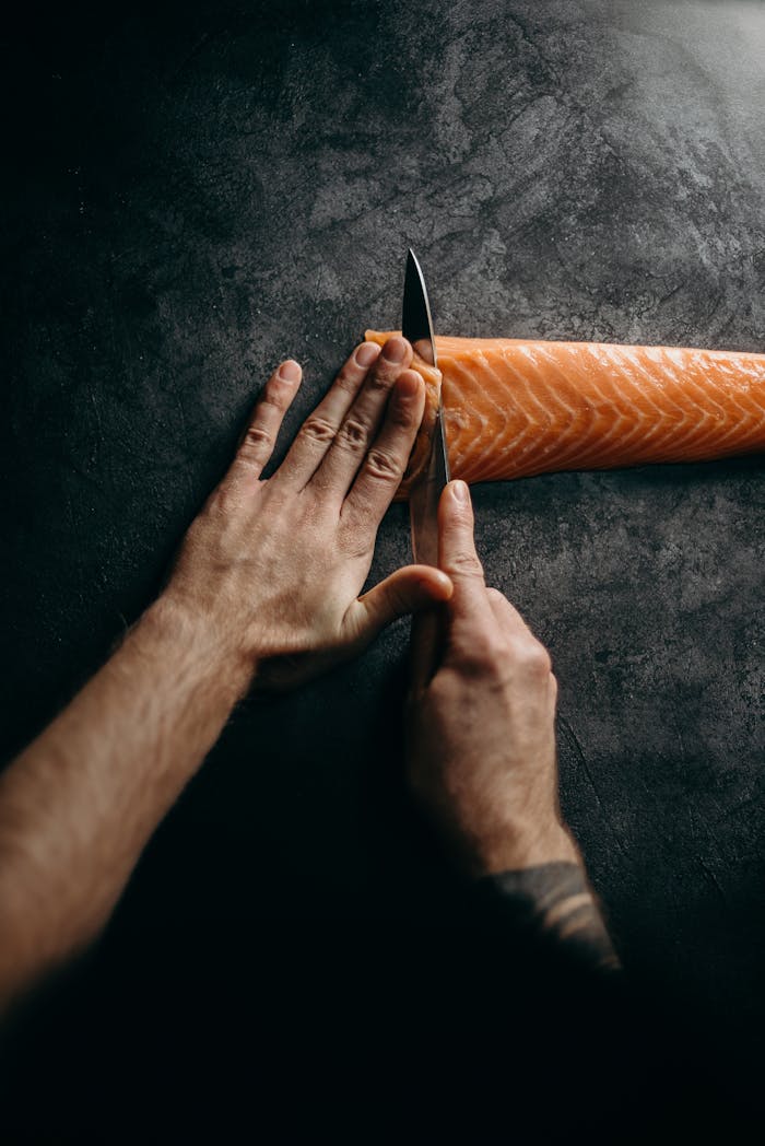 Chef expertly slicing fresh salmon on dark surface, highlighting culinary skill and precision in food preparation.